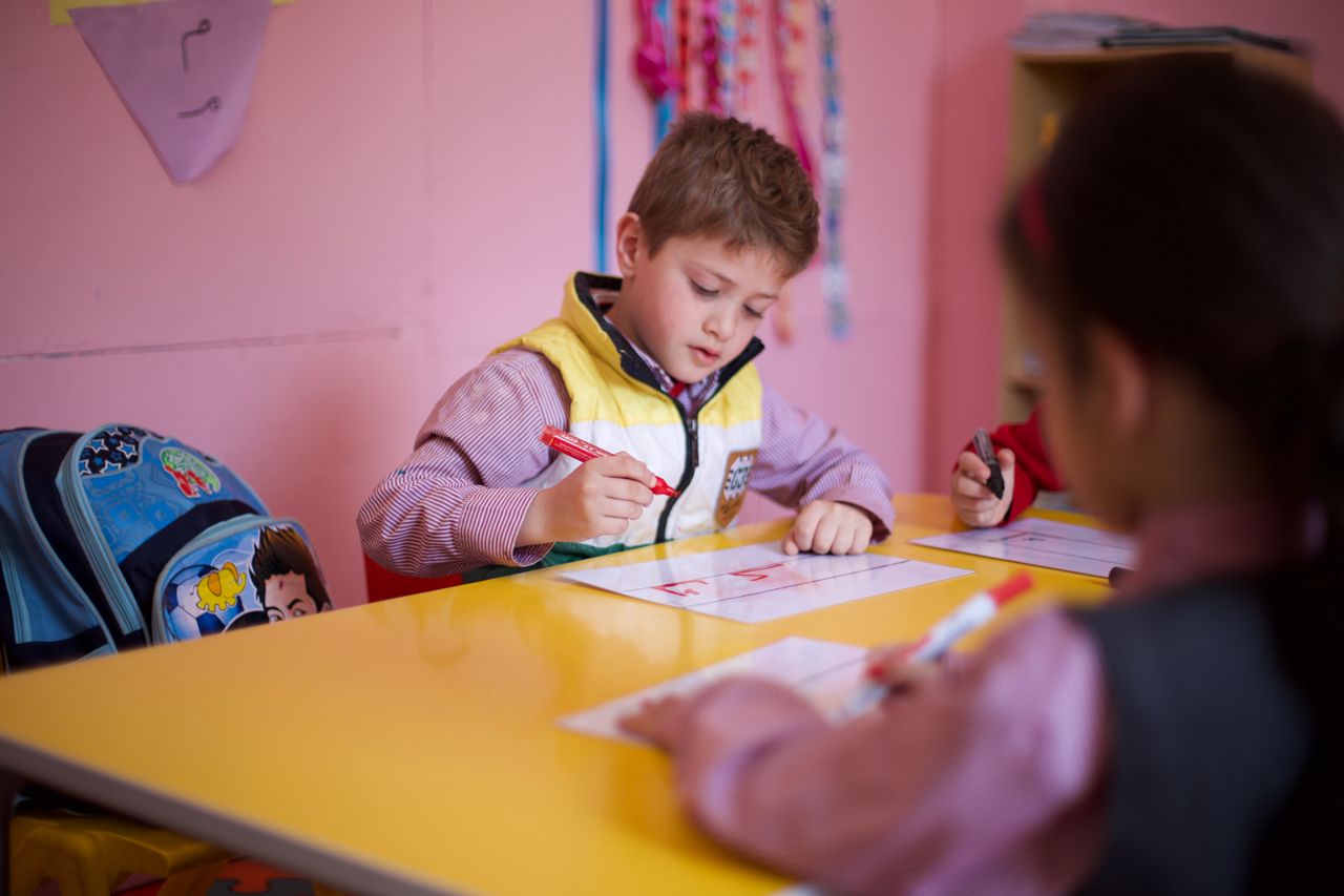 Children focused on drawing with markers inside a colorful preschool classroom setting.